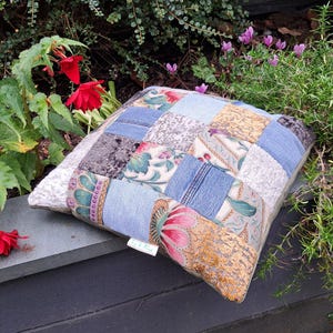 May include: A square patchwork cushion featuring a mix of fabrics, such as denim, floral prints, and textured materials. The cushion is positioned outdoors near plants and red flowers. A small tag with text is attached to the cushion.