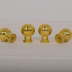 Könnte beinhalten: Vier goldfarbene Schrankknöpfe mit einer strukturierten, vertieften Oberfläche, die an Golfbälle erinnert. Die Knöpfe haben eine runde Form und einen kurzen, zylindrischen Sockel. Sie sind auf einer weißen Oberfläche angeordnet.