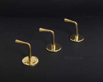 May include: Three gold metal shelf brackets with a curved arm and a round base. The brackets are arranged in a row on a black surface.