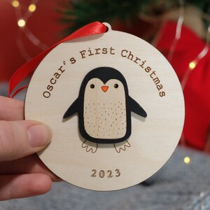May include: A wooden Christmas ornament with a penguin design. The ornament is round and features the text "Oscar's First Christmas" and the year "2023". The penguin is black and white with an orange beak. A red ribbon is attached for hanging.