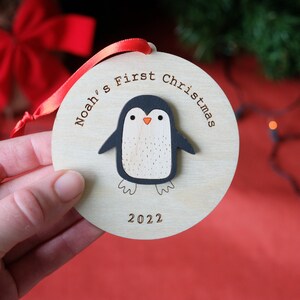 May include: Wooden Christmas ornament with a penguin design. The ornament is round with the text "Noah's First Christmas 2022". The penguin has a black body, white belly, and orange beak. A red ribbon is attached for hanging.