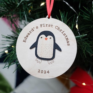 Wooden round bauble with red ribbon. Hand painted penguin with orange beak in the centre surrounded by your choice of engraved text. For example Edwards First Christmas 2024