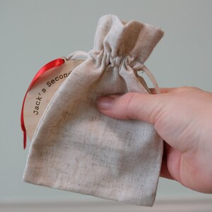 May include: A small, beige drawstring bag with a wooden tag attached by a red ribbon. The tag has the text "Jack's Second" printed on it. The bag is held in a person's hand.
