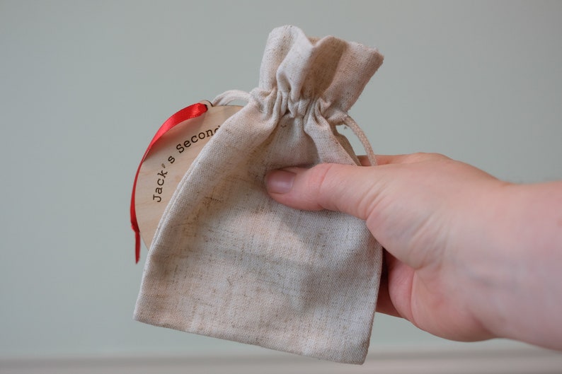 May include: A small, beige drawstring bag with a wooden tag attached by a red ribbon. The tag has the text "Jack's Second" printed on it. The bag is held in a person's hand.