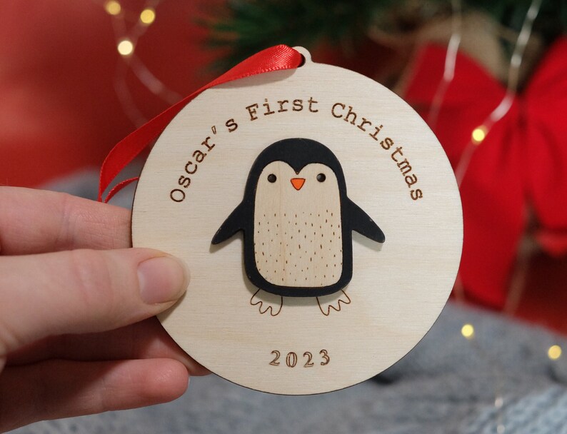 May include: A wooden Christmas ornament with a penguin design. The ornament is round and features the text "Oscar's First Christmas" and the year "2023". The penguin is black and white with an orange beak. A red ribbon is attached for hanging.