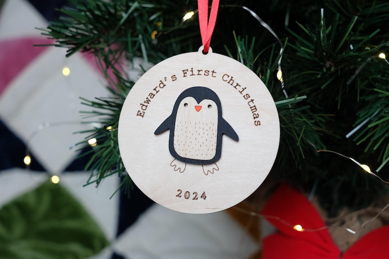 Wooden round bauble with red ribbon. Hand painted penguin with orange beak in the centre surrounded by your choice of engraved text. For example Edwards First Christmas 2024
