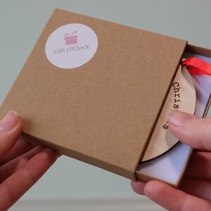 May include: A wooden Christmas ornament with the word "Christmas" printed on it, inside a brown box. The ornament has a red ribbon attached. The box has a "Gift O'Clock" sticker on the top.