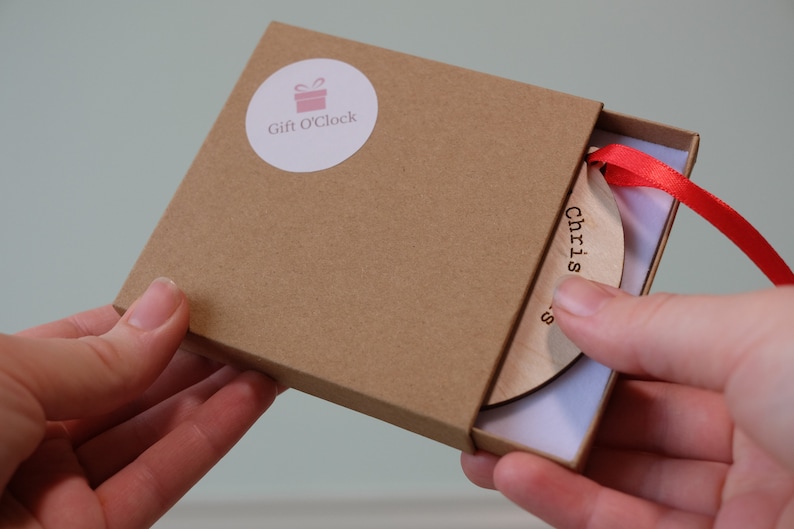 May include: A wooden Christmas ornament with the word "Christmas" printed on it, inside a brown box. The ornament has a red ribbon attached. The box has a "Gift O'Clock" sticker on the top.