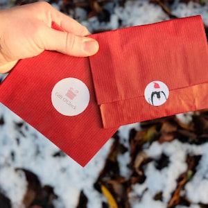 May include: Two red paper gift bags with white circular stickers. One sticker has the text "Gift O'Clock" and a pink gift box graphic. The other sticker features a penguin wearing a Santa hat. The bags are held in a hand.