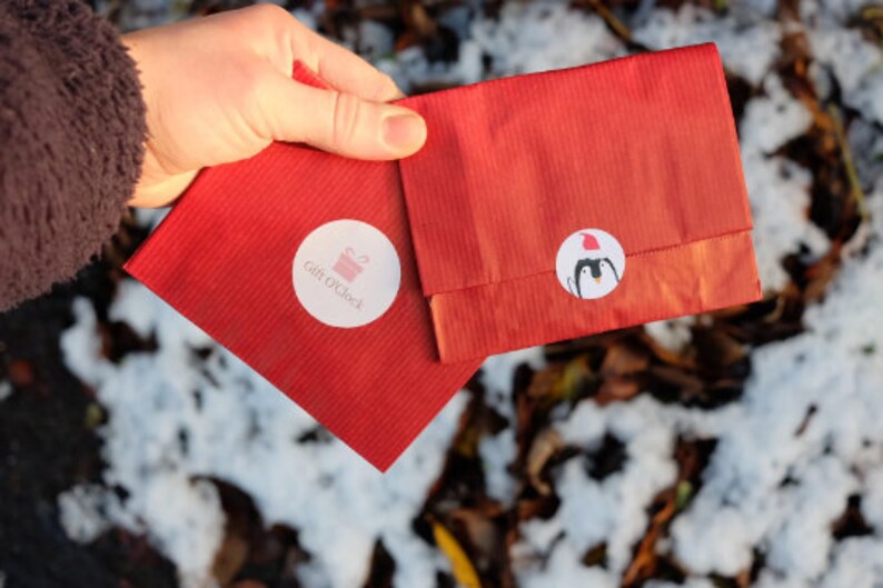 May include: Two red paper gift bags with white circular stickers. One sticker has the text "Gift O'Clock" and a pink gift box graphic. The other sticker features a penguin wearing a Santa hat. The bags are held in a hand.