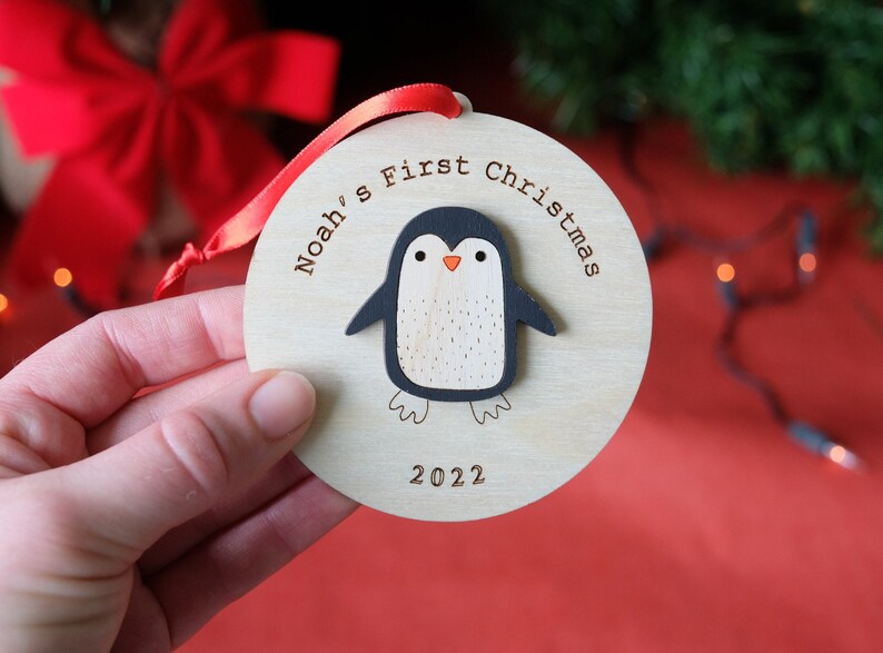 May include: Wooden Christmas ornament with a penguin design. The ornament is round with the text "Noah's First Christmas 2022". The penguin has a black body, white belly, and orange beak. A red ribbon is attached for hanging.