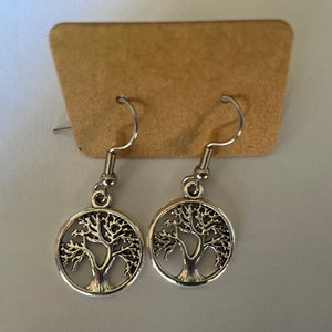 May include: A pair of silver dangle earrings featuring a tree of life design. The earrings are on a brown card.