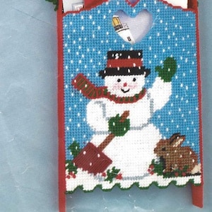 Puede incluir: Un muñeco de nieve sosteniendo una pala se encuentra frente a un conejo marrón. El muñeco de nieve lleva un sombrero rojo y negro y una bufanda verde. El muñeco de nieve y el conejo están sobre un fondo azul con puntos blancos. El fondo es un porta tarjetas con forma de trineo. El porta tarjetas es rojo y blanco con una abertura en forma de corazón. El texto "SNOWMAN SLED" está en la parte superior de la imagen. El texto "WALL DECORATION CARD HOLDER SIZE: 8x13 1/2" está debajo del muñeco de nieve.