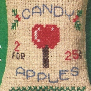 May include: A beige fabric bag with a green border and the words "CANDY 2 FOR 25¢ APPLES" embroidered in blue and red thread. The bag has a fringe border and a red apple design in the center.