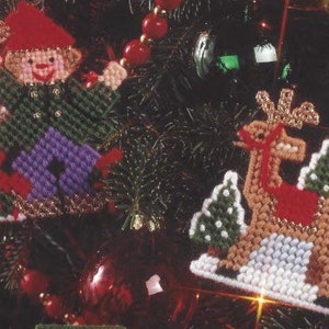 Puede incluir: Dos adornos navideños, uno de un elfo rojo y verde que sostiene un bastón de caramelo y el otro de un reno marrón con cuernos rojos de pie frente a dos árboles verdes.