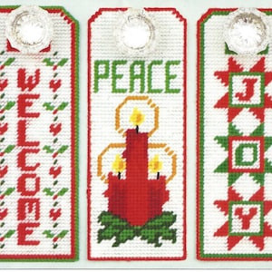 May include: Three white canvas door hangers with red and green embroidery. The first hanger says "WELCOME" in red letters. The second hanger has a red candle with yellow flames and says "PEACE" in green letters. The third hanger has a red and green patchwork design and says "JOY" in red letters.