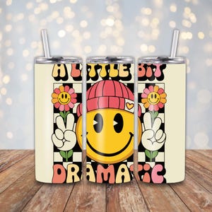May include: Three stainless steel tumblers with a retro design. Each tumbler features a yellow smiley face wearing a red beanie, peace signs, and the words "A Little Bit Dramatic" in a groovy font. The background is a checkerboard pattern.