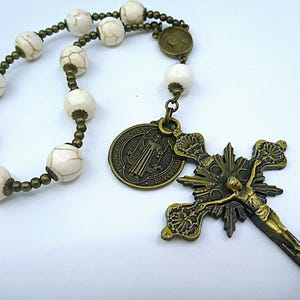 May include: A rosary with off-white, crackled beads and bronze-colored metal accents. The rosary features a crucifix with a detailed depiction of Jesus and a round pendant with religious imagery. The beads are approximately 0.5 inches in diameter.