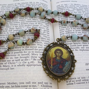 Pu&ograve; includere: Un rosario con perline azzurre, marroni e color crema. Il rosario presenta un ciondolo decorativo con un'immagine religiosa e una catena color bronzo. Le perline sono infilate su una catena color bronzo, appoggiate su un libro aperto.