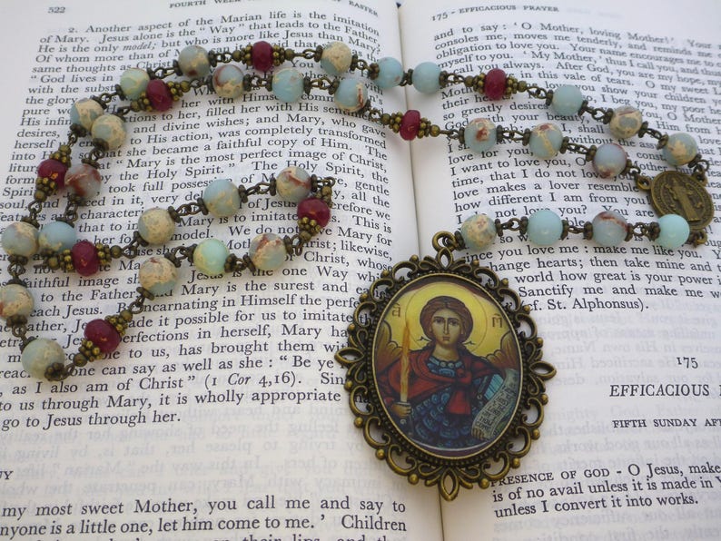 Pu&ograve; includere: Un rosario con perline azzurre, marroni e color crema. Il rosario presenta un ciondolo decorativo con un'immagine religiosa e una catena color bronzo. Le perline sono infilate su una catena color bronzo, appoggiate su un libro aperto.