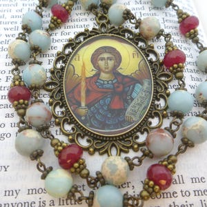 Pu&ograve; includere: Un rosario con un pendente centrale raffigurante l'immagine di un santo, circondato da una cornice decorativa in bronzo. Le perline del rosario sono un mix di perline blu, rosse e macchiate, collegate da catene di bronzo.