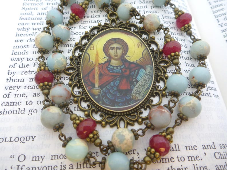 Pu&ograve; includere: Un rosario con un pendente centrale raffigurante l'immagine di un santo, circondato da una cornice decorativa in bronzo. Le perline del rosario sono un mix di perline blu, rosse e macchiate, collegate da catene di bronzo.