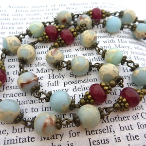Pu&ograve; includere: Una collana rosario con perline azzurre, bordeaux e beige. Le perline sono collegate da una catena color bronzo. Il rosario &egrave; appoggiato su un libro aperto con testo religioso.