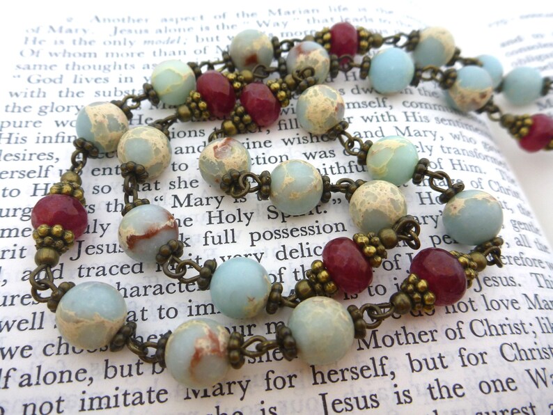 Pu&ograve; includere: Una collana rosario con perline azzurre, bordeaux e beige. Le perline sono collegate da una catena color bronzo. Il rosario &egrave; appoggiato su un libro aperto con testo religioso.