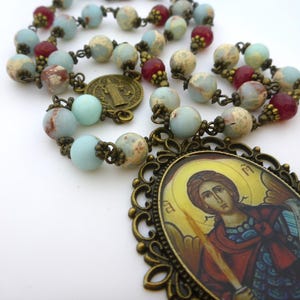 Pu&ograve; includere: Un rosario con perline blu, rosse e beige. Le perline sono infilate con metallo color bronzo. Un ciondolo ovale color bronzo con l'immagine di un santo si trova nella parte inferiore del rosario.