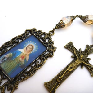 May include: A gold-toned rosary with a rectangular pendant featuring a blue and white image of the Virgin Mary. The pendant is decorated with ornate floral designs. The rosary has clear glass beads and a crucifix with a figure of Jesus.