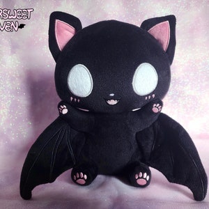 Puede incluir: Un murciélago de peluche negro con orejas y patas rosas. El murciélago tiene grandes ojos blancos y una pequeña boca rosa. El murciélago está sentado sobre un fondo rosa brillante.