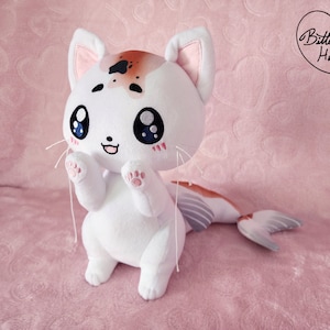 May include: White plush cat toy with a brown and orange fishtail. The cat has large blue eyes and a pink nose. The toy is sitting on a pink blanket with the text "Bittersweet Haven" in a heart.
