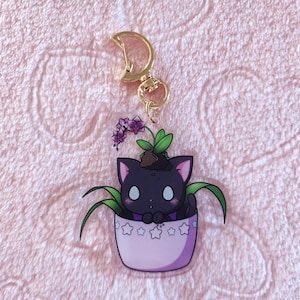 May include: A black cat with white eyes is peeking out of a pink flower pot with white stars. The cat is surrounded by green leaves and purple flowers. The pot is attached to a gold key chain.