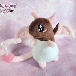 May include: A small, brown and white plush dragon with pink wings and a pink tail. The dragon has a heart-shaped charm attached to its tail.