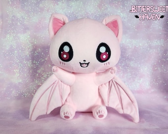May include: Pink plush bat with large eyes and a smiling mouth. The bat has large, pink wings with stitching details. The plush toy is sitting on a pink sparkly background.  Bittersweet Haven.