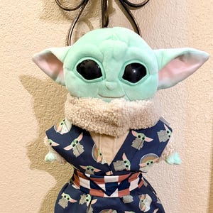 May include: A plush Grogu doll wearing a navy blue kimono-style outfit. The outfit is patterned with Grogu's likeness in white and gray. The doll has large black eyes, green skin, and large ears. The outfit has a white and brown belt.