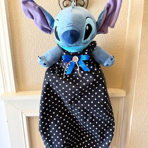 May include: A plush blue character with large ears and a black and white polka dot dress, adorned with a blue bow. The character is hanging from a decorative metal hook.