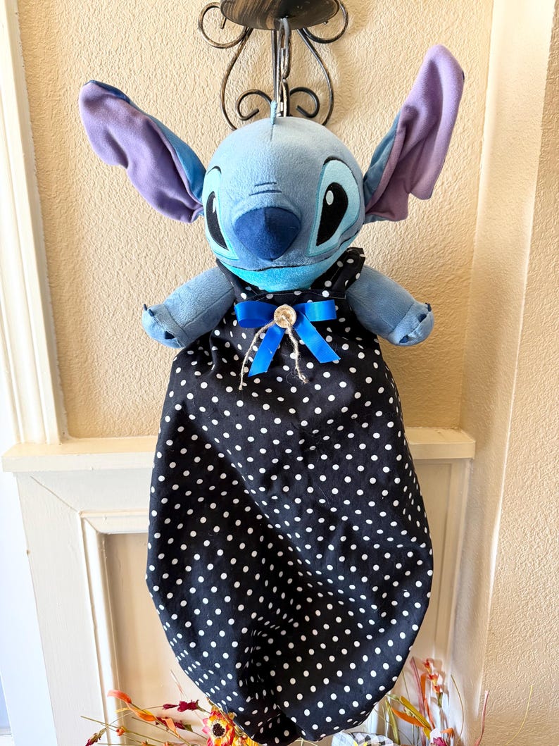 May include: A plush blue character with large ears and a black and white polka dot dress, adorned with a blue bow. The character is hanging from a decorative metal hook.