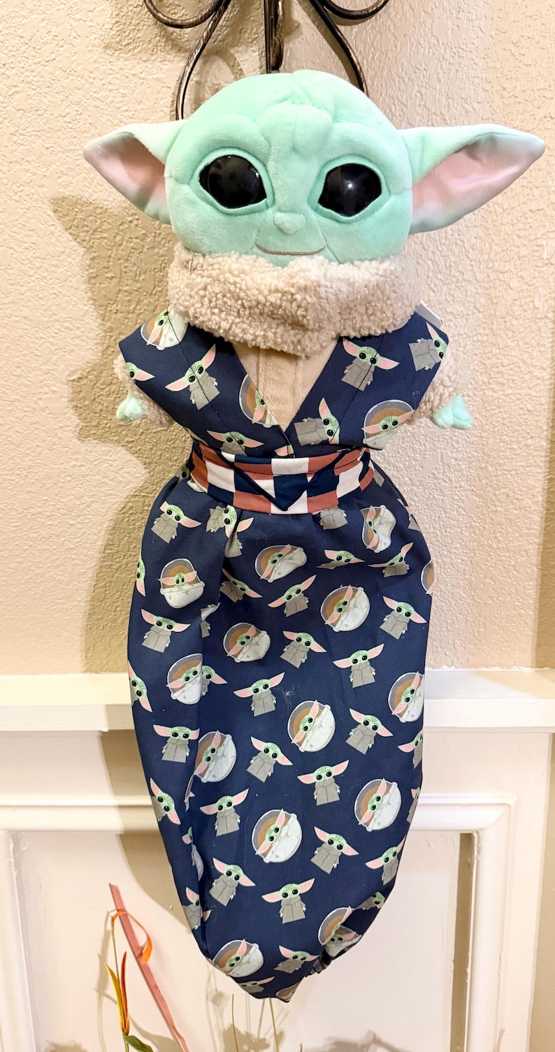 May include: A plush Grogu doll wearing a navy blue kimono-style outfit. The outfit is patterned with Grogu's likeness in white and gray. The doll has large black eyes, green skin, and large ears. The outfit has a white and brown belt.