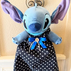 May include: A plush blue Stitch doll with large ears and a black and white polka dot dress. The dress is accented with a blue bow and a button. The doll is hanging from a decorative metal fixture.