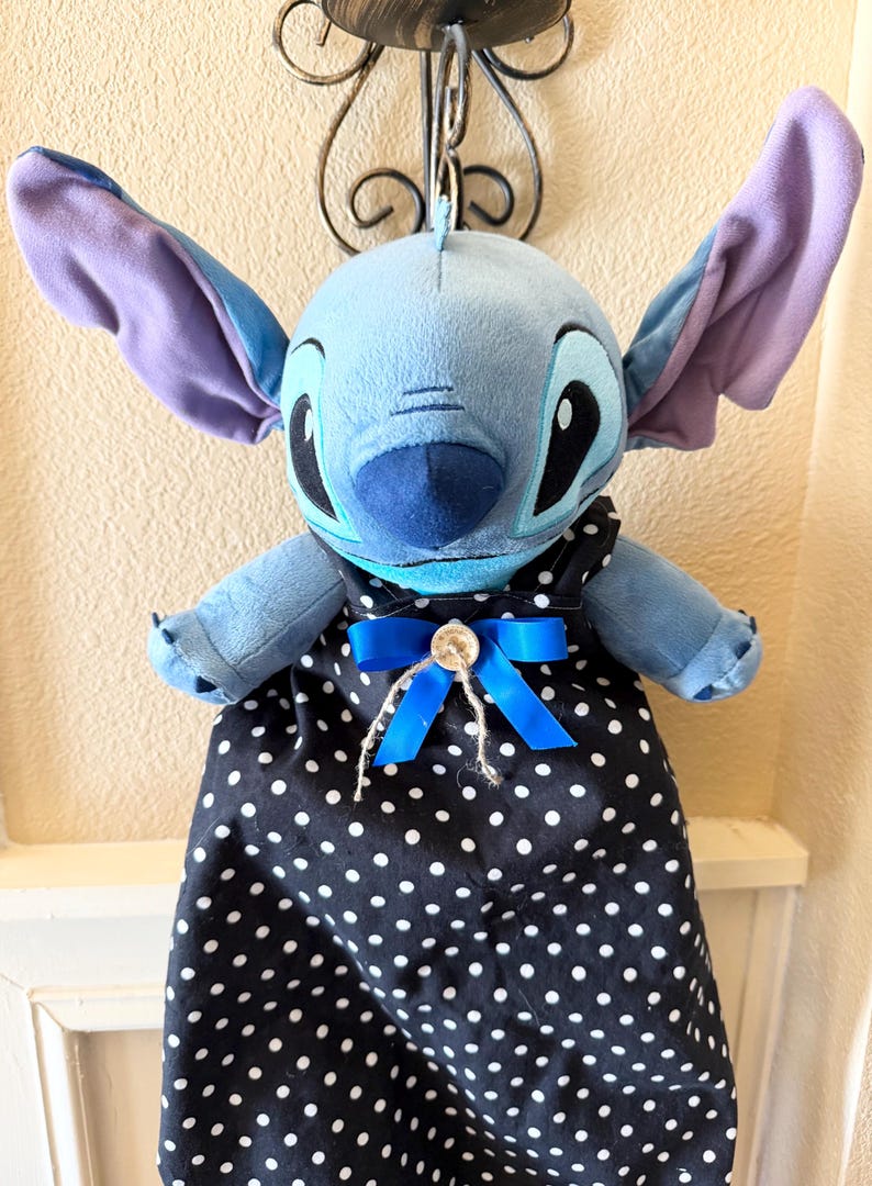 May include: A plush blue Stitch doll with large ears and a black and white polka dot dress. The dress is accented with a blue bow and a button. The doll is hanging from a decorative metal fixture.