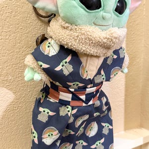 May include: A plush Grogu doll, also known as Baby Yoda, wearing a navy blue kimono-style outfit. The outfit is patterned with Grogu's likeness. The doll has large black eyes, large ears, and a tan fur collar.
