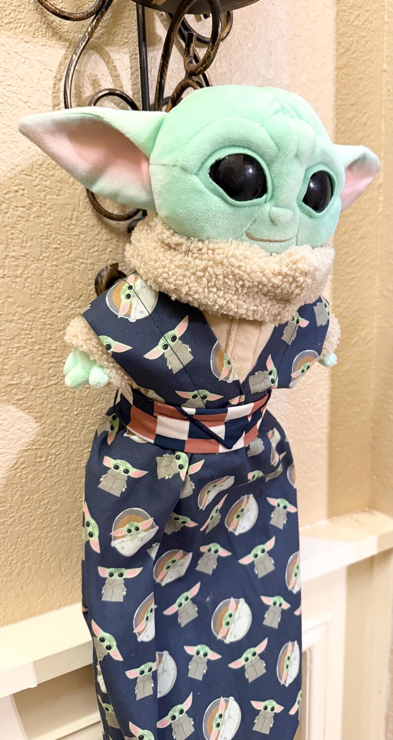 May include: A plush Grogu doll, also known as Baby Yoda, wearing a navy blue kimono-style outfit. The outfit is patterned with Grogu's likeness. The doll has large black eyes, large ears, and a tan fur collar.