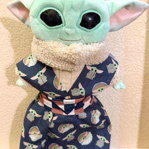 May include: A plush Grogu doll, also known as Baby Yoda, wearing a dark blue patterned robe. The robe features a repeating pattern of Grogu figures. The doll has large black eyes, large ears, and a light green complexion.