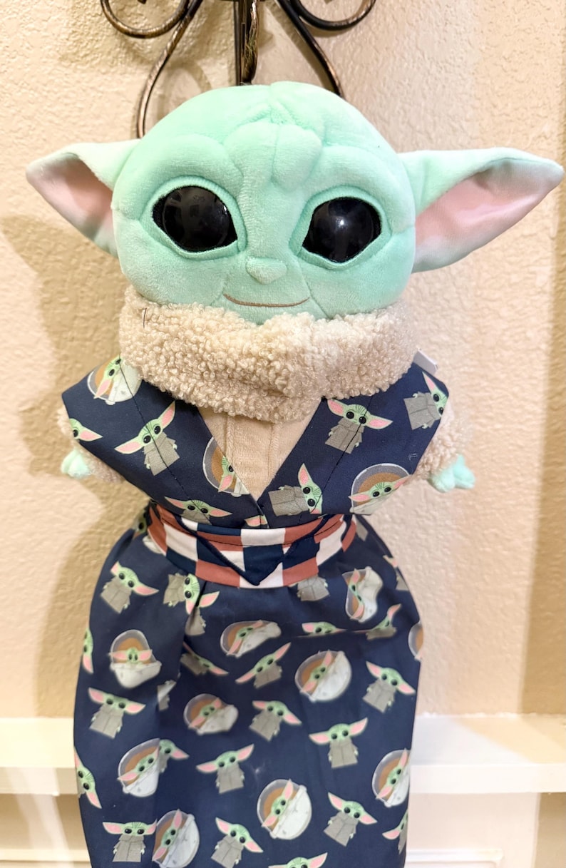 May include: A plush Grogu doll, also known as Baby Yoda, wearing a dark blue patterned robe. The robe features a repeating pattern of Grogu figures. The doll has large black eyes, large ears, and a light green complexion.
