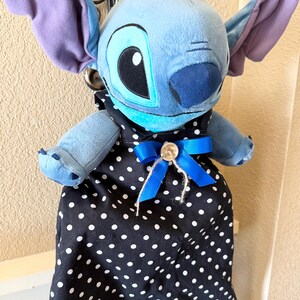 May include: A plush Stitch doll with large ears and a blue body, wearing a black apron with white polka dots. The apron is adorned with a blue bow and a button.