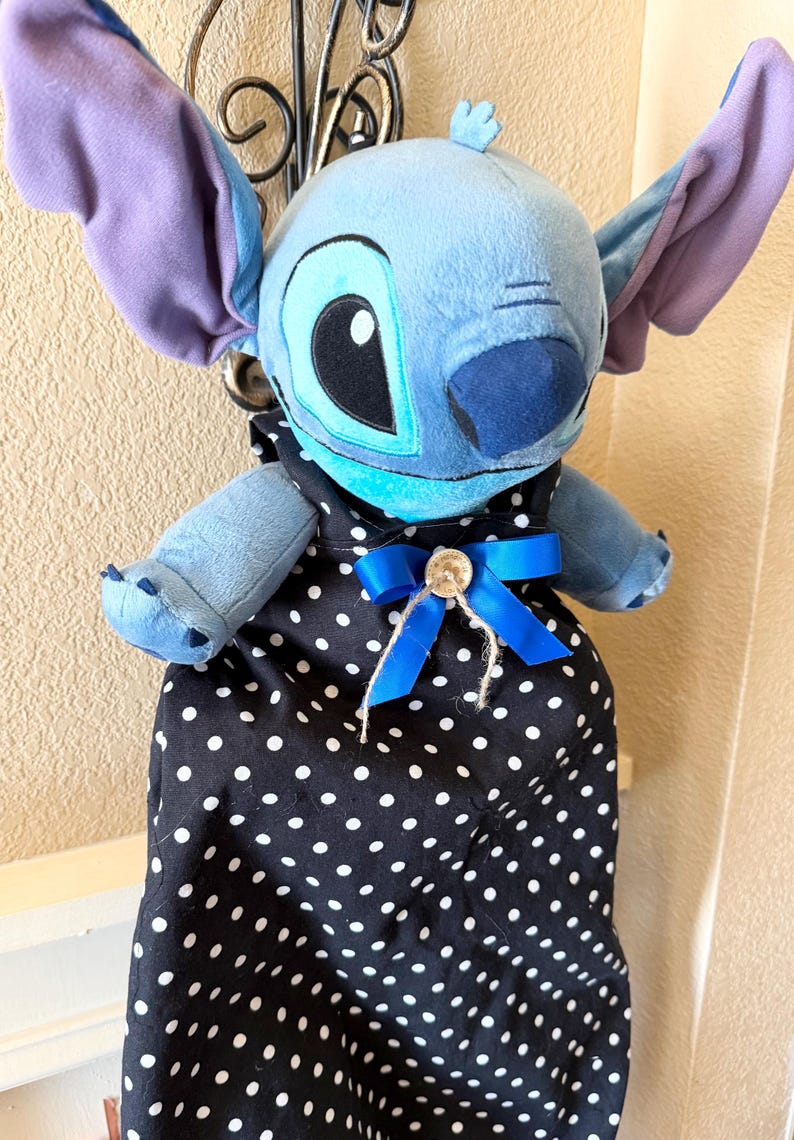 May include: A plush Stitch doll with large ears and a blue body, wearing a black apron with white polka dots. The apron is adorned with a blue bow and a button.