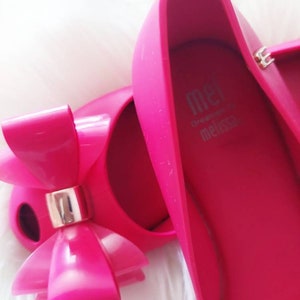 May include: A pair of bright pink ballet flats with a large pink bow on the toe. The shoes are made by Melissa and have the brand name on the inside of the shoe.