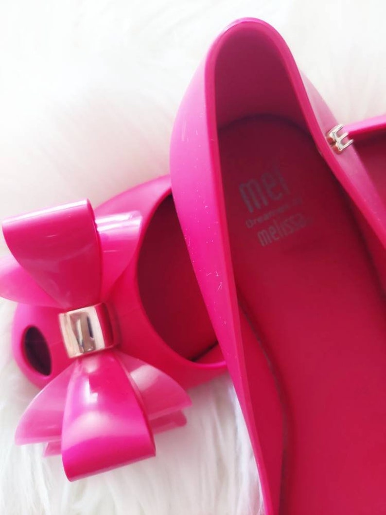 May include: A pair of bright pink ballet flats with a large pink bow on the toe. The shoes are made by Melissa and have the brand name on the inside of the shoe.