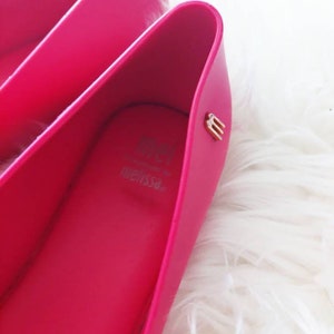 May include: A pair of bright pink ballet flats with a gold "M" logo on the inside of the shoe. The shoes are made of a rubber-like material and have a simple, sleek design.